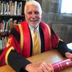 Dr Keith Hiscock, smiling, with white hair and beard, in a black and red ceremonial academic gown, white shirt and yellow striped tie, sits at a large wooden desk holding a red ceremonial scroll with gold lettering, with a large bookcase full of books in the background.