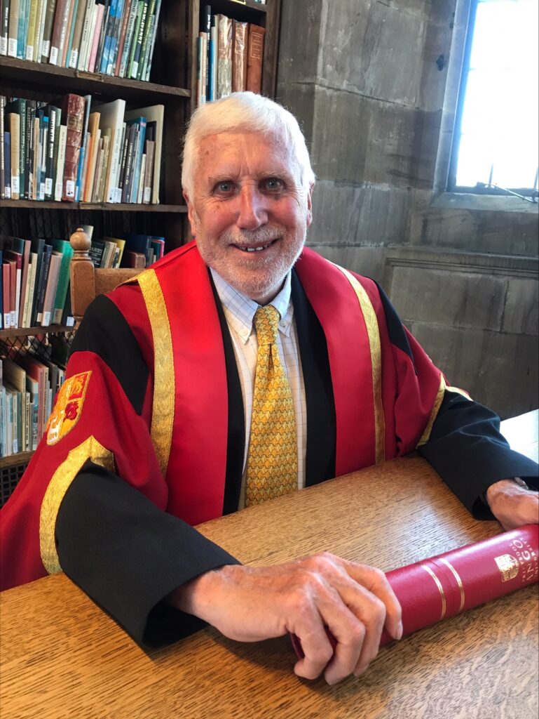 Dr Keith Hiscock, smiling, with white hair and beard, in a black and red ceremonial academic gown, white shirt and yellow striped tie, sits at a large wooden desk holding a red ceremonial scroll with gold lettering, with a large bookcase full of books in the background.